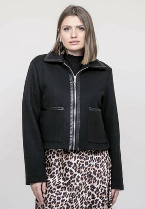 Femme aux cheveux châtain clair raides portant une veste noire zippée avec des garnitures en cuir et une jupe imprimée léopard, debout devant un fond uni.