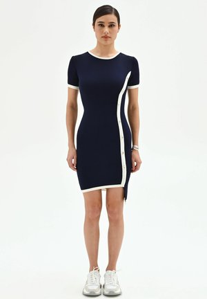 Femme portant une robe ajustée bleu marine à manches courtes avec des bordures blanches et des boutons, associée à des baskets blanches, debout devant un fond blanc uni.