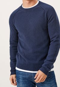 Homme portant un pull en maille bleu marine par-dessus une chemise blanche, associé à un jean bleu, debout devant un fond uni.