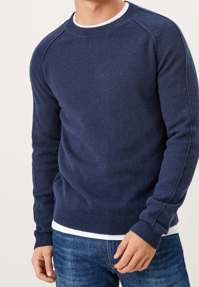 Homme portant un pull en maille bleu marine par-dessus une chemise blanche, associé à un jean bleu, debout devant un fond uni.