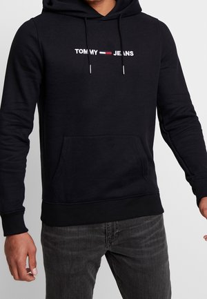 Czarna bluza z kapturem z białym napisem „Tommy Jeans” oraz logo w kolorach czerwonym, białym i granatowym na piersi, noszona z ciemnoszarymi dżinsami na stojącej osobie.