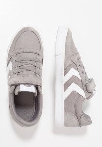 Zapatillas infantiles grises con rayas en forma de chevron blancas, cordones y tiras de velcro mostradas desde arriba y de lado sobre un fondo blanco.