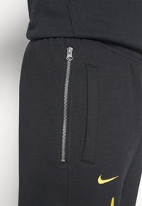 Sorte træningsbukser med en lynlåslomme og et broderet gult Nike-logo. Stoffet ser blødt ud med en glat tekstur.