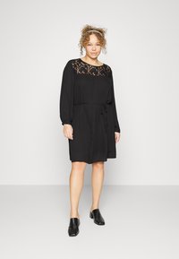 Vestido preto com decote de renda, mangas longas e faixa na cintura. Tecido suave, até ao joelho, combinado com mules pretas de salto.