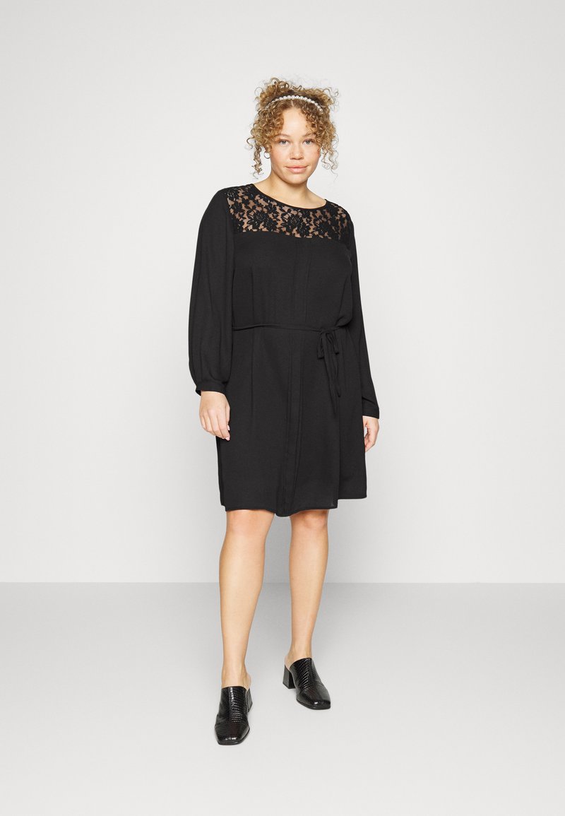 Vestido preto com decote de renda, mangas longas e faixa na cintura. Tecido suave, até ao joelho, combinado com mules pretas de salto.