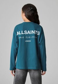 Tirkizna majica dugih rukava s bijelim tiskanim tekstom na leđima, uključujući "ALLSAINTS" i "LONDON," s okruglom izrezom i opuštenim krojem.