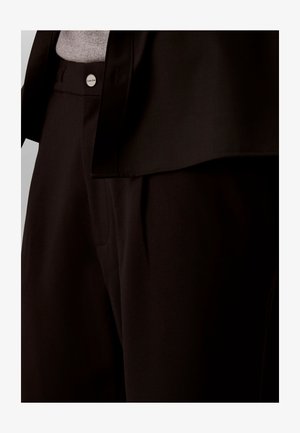 Nahaufnahme einer Person, die schwarze Hosen mit sichtbarem Knopf trägt, sowie einen passenden schwarzen Blazer über einem grauen Oberteil.