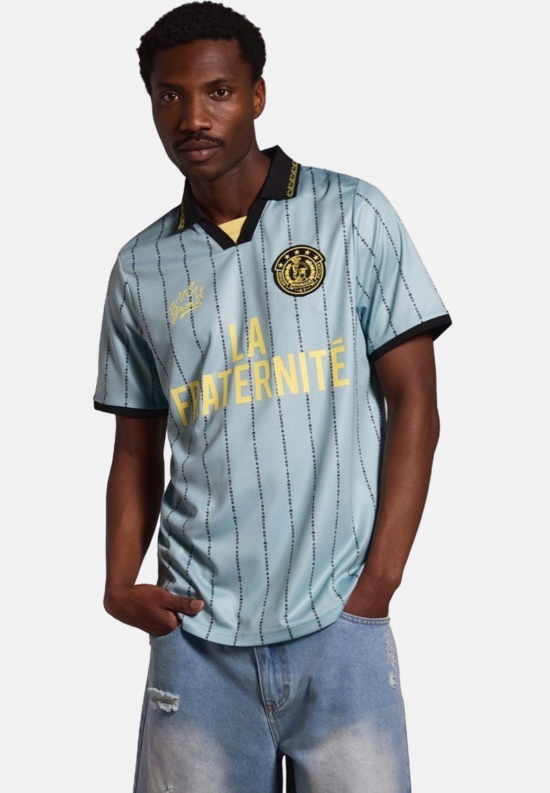 Jersey de manga corta azul claro con rayas verticales sutiles, cuello negro y texto amarillo "LA FRATERNITÉ." Presenta un emblema en el pecho.