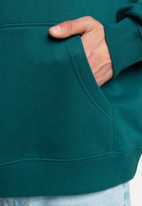 Tyrkysová mikina s rebrovanými manžetami a lemom. Obsahuje bočnú kapsu; látka vyzerá jemne a textúrovane. Zamerajte sa na dizajn vrecka a farbu.