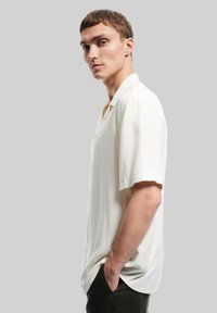 Camisa de manga corta, color crema, con un corte relajado, hecha de un tejido ligero, que presenta un cuello abotonado y un brillo sutil.