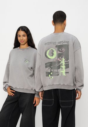 Deux personnes portent des sweats gris avec des motifs graphiques abstraits, l'une fait face, l'autre montre un grand motif imprimé dans le dos.