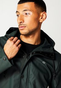 Dark green waterproof jacket with a zip closure, hood, and adjustable drawstrings; features a logo accent and smooth, glossy texture.