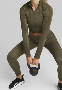 Set sportivo di un verde oliva scuro con una maglia a maniche lunghe con zip e un motivo testurizzato, abbinato a leggings coordinati e un kettlebell nero.