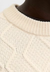 Grosser Creme-Pullover mit einem gerippten Kragen, der ein strukturiertes Rautenmuster auf einer gestrickten Oberfläche zeigt. Dunkler innerer Kragen sichtbar.