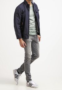 Marineblauwe lichte jas met een staande kraag, grijs T-shirt, lichtgrijze jeans en witte sneakers met zwarte strepen. Casual stedelijke outfit.