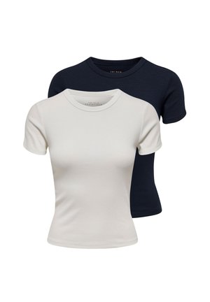 Jeune femme portant un haut blanc à manches courtes, un jean bleu taille haute, des talons noirs et des colliers dorés, debout avec la main sur la hanche sur fond blanc.