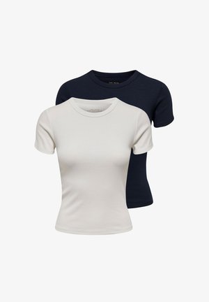 Jeune femme portant un haut blanc à manches courtes, un jean bleu taille haute, des talons noirs et des colliers dorés, debout avec la main sur la hanche sur fond blanc.
