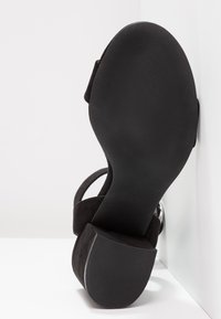 Sandale noire à semelle plate et talon bloc, vue de dessous sur fond blanc en coin.