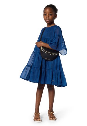 Robe bleue à volants avec des manches transparentes évasées et un motif de feuilles. Sac bandoulière noir orné de clous dorés. Portée avec des sandales marron.