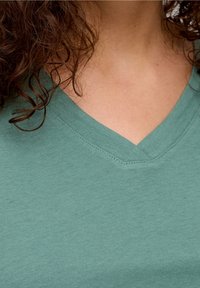 Capelli ricci scuri sulle spalle, indossando una maglietta a V color teal, che mostra il collo e la parte superiore del torace.
