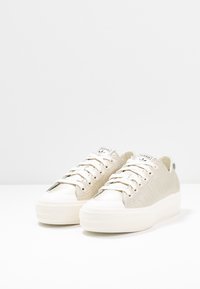 Béžové tenisky s hladkým koženým svrškem, zaoblenou špičkou a bílou gumovou podrážkou. Mají zavazování na tkaničky a tři boční pruhy.