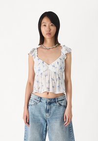 Floral cropped top with ruffled straps, featuring a light cream color with blue flowers. Paired with loose denim jeans.