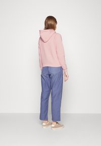 Sudadera de algodón rosa con un bolsillo tipo canguro, combinada con pantalones de corte relajado azul y zapatillas beige, vista desde la parte de atrás.