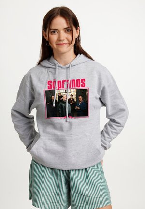 Młoda kobieta z długimi brązowymi włosami nosi szarą bluzę z kapturem z zdjęciem obsady serialu The Sopranos i czerwonym napisem, do tego zielone i białe krótkie spodenki w paski.