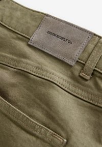 Olive roheline puuvillane kangas, millel on peene tekstuur, koos õmmeldud sildiga, millel on kirjas "DENIM SUPPLY Co." ja tagatasku disain.