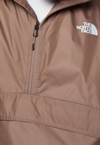 Brązowy kurtka przeciwwiatrowa z pół-zamkiem, wykonana z lekkiego materiału, z białym logo marki na lewej piersi. Widoczne płaskie szwy.