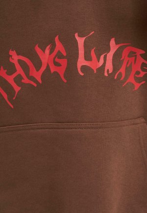 Pruun kapuutsiga, mille esiosas on suur punane graafiline tekst "thug life" stiliseeritud, kiharates kirjas.