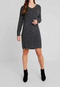 Robe en maille grise avec col en V, manches longues et coupe droite. Texture douce, ourlet arrivant à mi-cuisse, associée à des bottines noires.