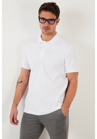 Camisa polo blanca de canalé con cuello estructurado y tres botones, combinada con pantalones grises de rayas. Presenta mangas cortas y un ajuste relajado.
