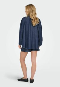 Nainen, jolla on pitkät vaaleat hiukset, käyttää tummansinistä pitkähihaista paitaa ja siihen sopivia shortseja, seisoo selin mustat loafersit jalassa.