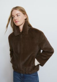 Chaqueta de piel sintética marrón con cuello alto, cierre de cremallera, dobladillo acanalado y bolsillos laterales. Textura suave con un ajuste relajado.