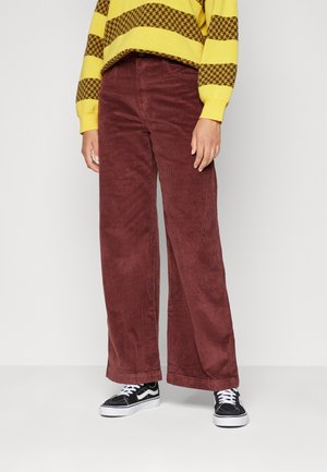 Pantalons à taille large en velours côtelé de couleur bourgogne foncé, présentant un motif côtelé texturé, associés à des baskets noires et un pull à rayures jaunes.