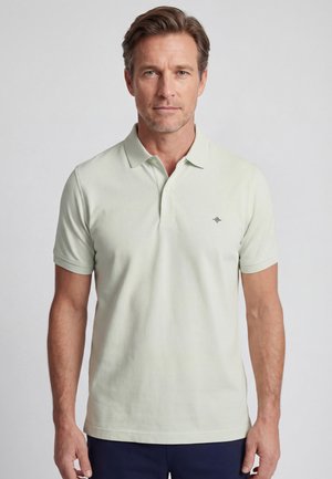 Man die een lichtgroen poloshirt met korte mouwen en een klein logo op de borst draagt, staande tegen een effen lichtgrijze achtergrond.
