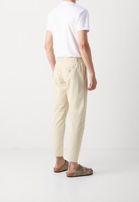 Mężczyzna w beżowych, taperowanych spodniach z elastycznym pasem, białej koszuli z krótkim rękawem i brązowych zamszowych sandałach na klamerkę, stojący na białej podłodze.