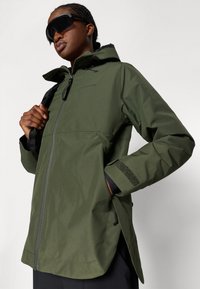 Casaco verde impermeável com fecho zip, capuz ajustável e detalhes com fecho de Velcro. Apresenta um corte relaxado e bolsos laterais.