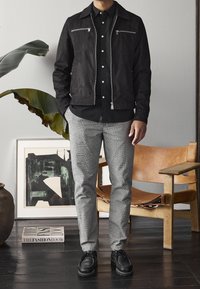 Zwarte suède jas met zilveren ritssluitingen over een zwart shirt; grijs geruite broek; zwarte schoenen; binnenomgeving met planten en kunst op de achtergrond.