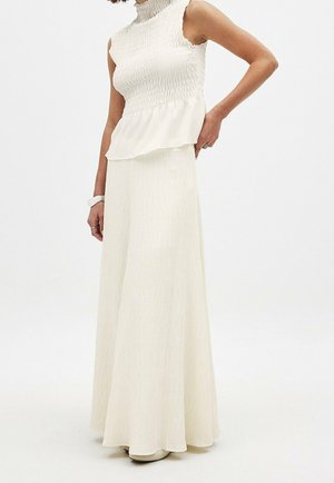 Długa kremowa sukienka z górą w stylu smock, bez rękawów i zwiewnym, teksturowanym dołem. Materiał ma subtelny połysk i delikatny, lekki wygląd.