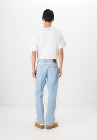 Tricou alb cu mânecă scurtă și branding albastru pe spate, asociat cu blugi drepti de culoare albastru deschis și pantofi slip-on bej.