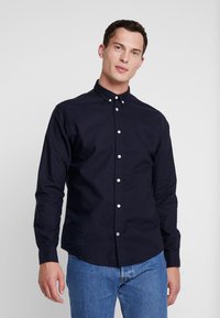 Camisa de botones azul marino de algodón, con cuello, botones blancos y un ligero brillo, combinada con jeans azul claro.