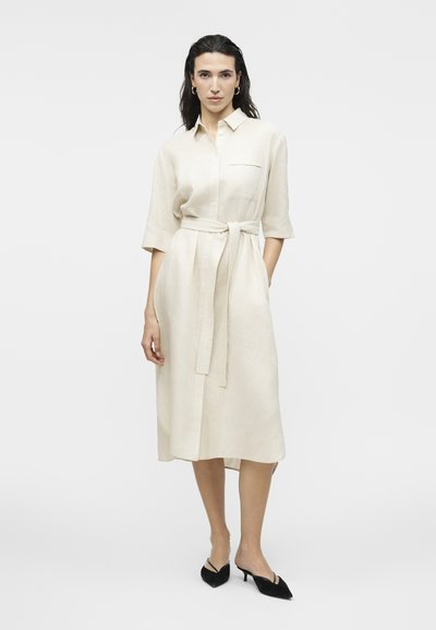 Γυναίκα με σκούρα μαλλιά φοράει μπεζ midi φόρεμα με κουμπιά και ζώνη στη μέση, καθώς και μαύρα μυτερά παπούτσια με τακούνι kitten, στέκεται μπροστά σε απλό φόντο.