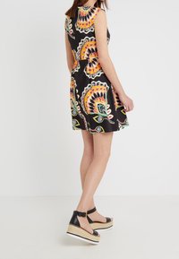 Musta mekko värikkäällä paisley-kuviolla, olkaimeton malli, vartalonmyötäinen vyötärö ja levenevä hame. Mustat korkeat sandaalit nilkkaremmillä.