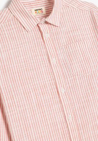 Lichtgewicht kindershirt met verticale roze en witte strepen, knoopsluiting aan de voorkant en een enkele borstzak. Katoenen stof.