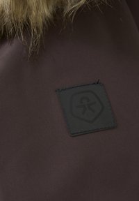 Brunt tyg med en texturerad yta har en svart rektangulär etikett präglad med en logotyp. Designen inkluderar en faux pälsdetalj på toppen.