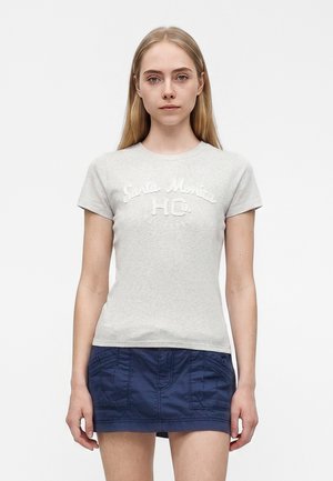 Junge Frau mit glatten blonden Haaren, die ein hellgraues "Santa Monica" T-Shirt und einen marineblauen Minirock trägt, steht vor einem weißen Hintergrund.