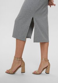 Beige patent leather heeled sandals with a block heel, open toe, and adjustable ankle strap. The model wears a gray ribbed midi dress.