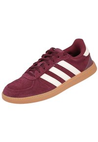 Zapatilla de gamuza color marrón con diseño de tres rayas blancas, puntera redonda, cierre de cordones en la parte delantera, cuello acolchado y suela de goma.